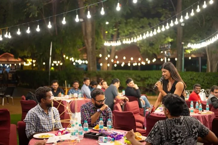 A host with a microphone addressing colleagues at an outdoor company dinner under string lights